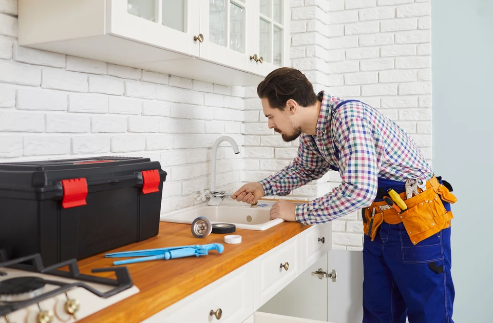 Un plombier en salopette bleue et chemise à carreaux répare un robinet de cuisine dans un intérieur moderne avec mur en briques blanches