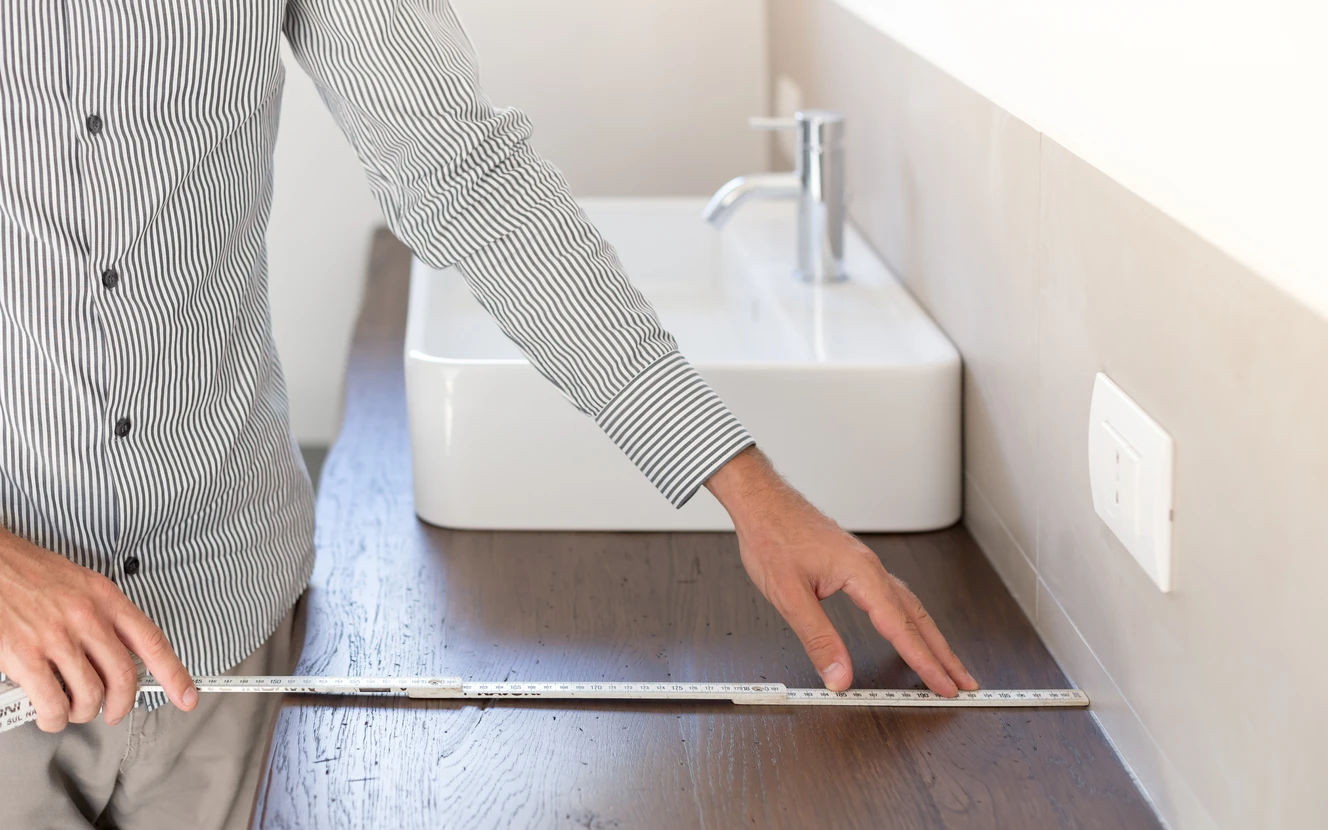 Une personne en chemise rayée mesure avec une règle métallique graduée l'espace entre un lavabo moderne blanc et un mur dans une salle de bain contemporaine aux tons beiges