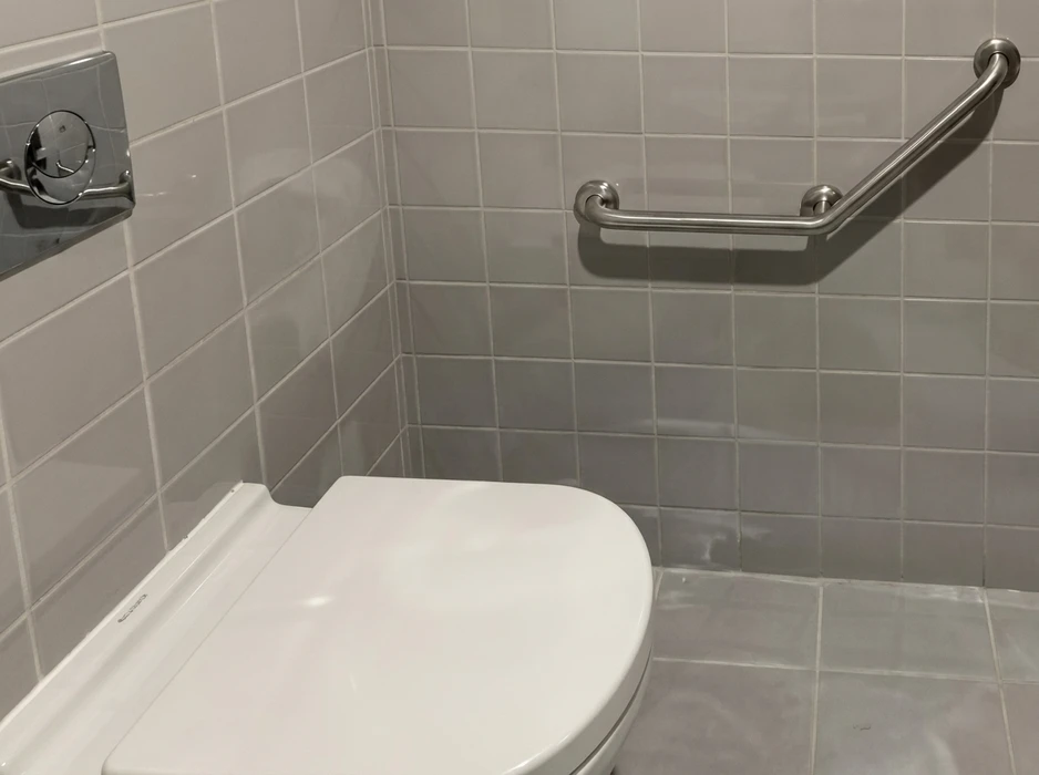 Salle de bains adaptée PMR avec WC suspendu blanc, carrelage mural et sol gris, barre d'appui en inox coudée fixée au mur, et chasse d'eau encastrée à déclenchement pneumatique