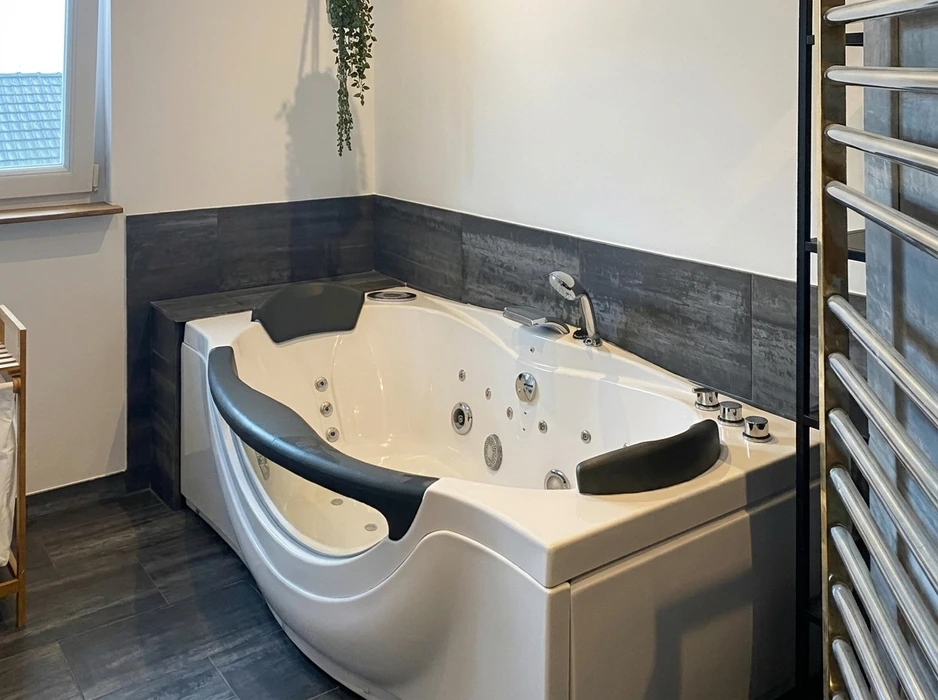 Salle de bain moderne avec baignoire balnéo d'angle biplace blanche, équipée de buses de hydrothérapie, deux appuie-têtes ergonomiques noirs, robinetterie chromée centrale, bordure en carrelage imitation bois gris anthracite