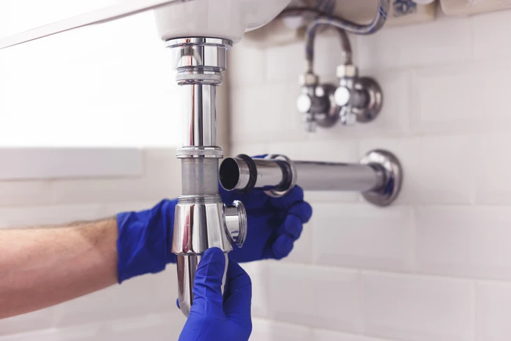 Plombier professionnel en uniforme bleu travaillant sous un évier de cuisine, manipulant la tuyauterie d'évacuation