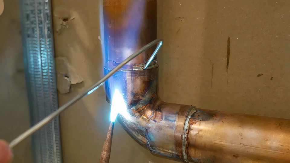 Un plombier en veste rouge utilise un chalumeau à gaz pour souder une canalisation en cuivre dans un environnement intérieur